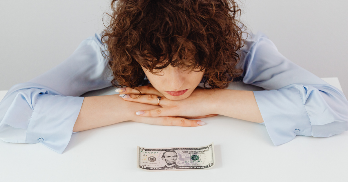 Woman resting her chin on her hands, looking down at a single five-dollar bill on a white table