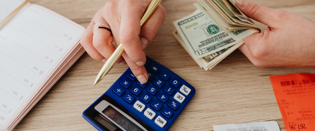 Hands calculating finances with a calculator, cash, and planner on a desk.