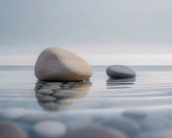 Two smooth stones resting in calm, still water with soft reflections, against a hazy blue-gray sky.