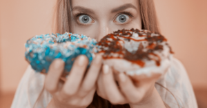 Woman holding two donuts up to her eyes, one blue-sprinkled and one chocolate-drizzled, with a wide-eyed expression.