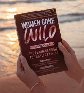 Hands holding the book 'Women Gone Wild: Leadership – The Feminine Guide to Fearless Living' by Rhonda Swan and Jules Schroeder, against a beach sunset backdrop.