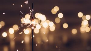 A lit sparkler emits bright white sparks against a dark background filled with warm, golden bokeh lights, creating a romantic, celebratory glow.