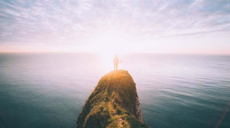 A person standing on a cliff jutting out into the ocean with the sunrise in front of them