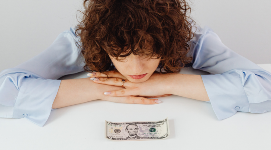 Woman resting her chin on her hands, looking down at a single five-dollar bill on a white table